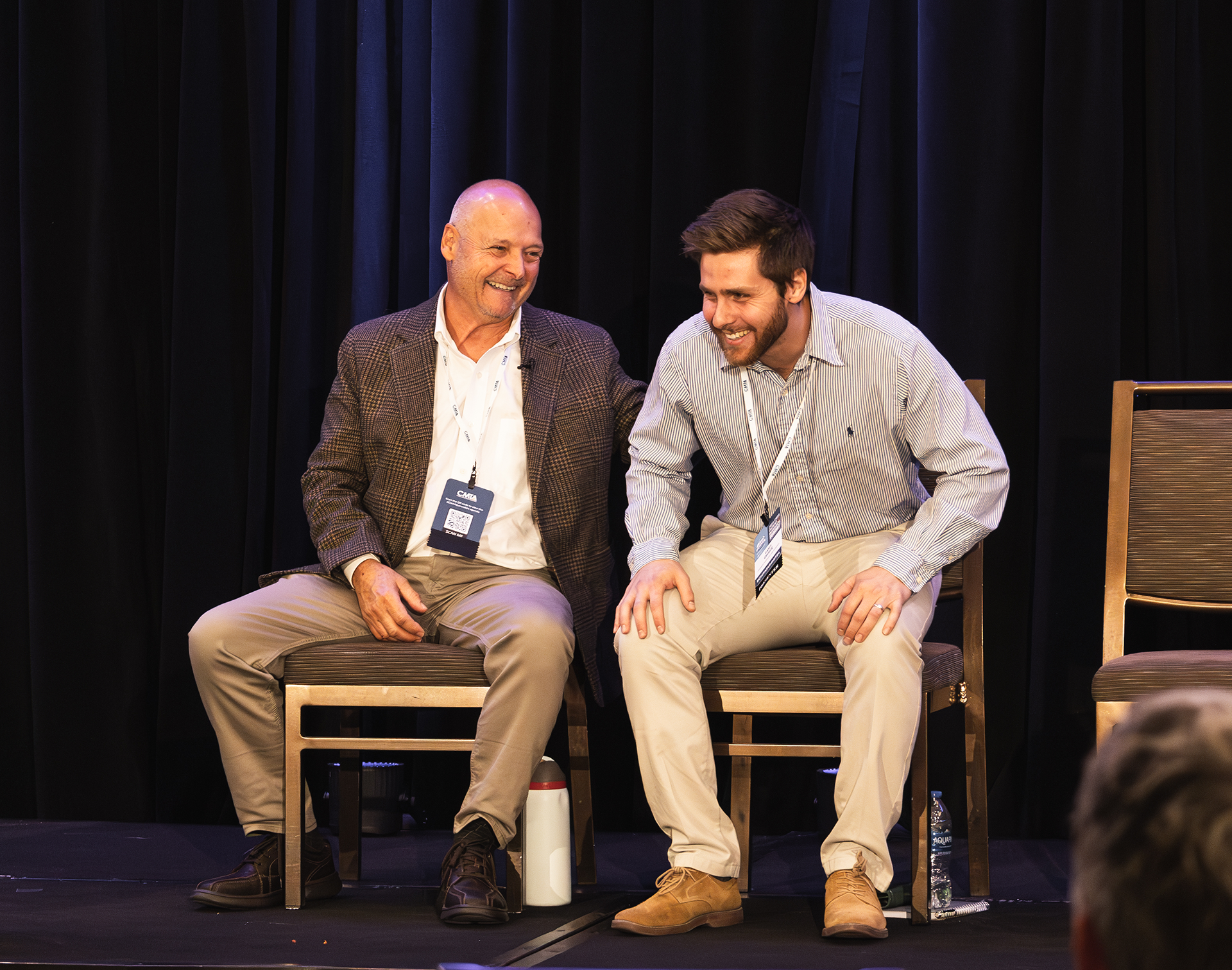 Ken Cornell and another man sitting in chairs laughing on a stage.